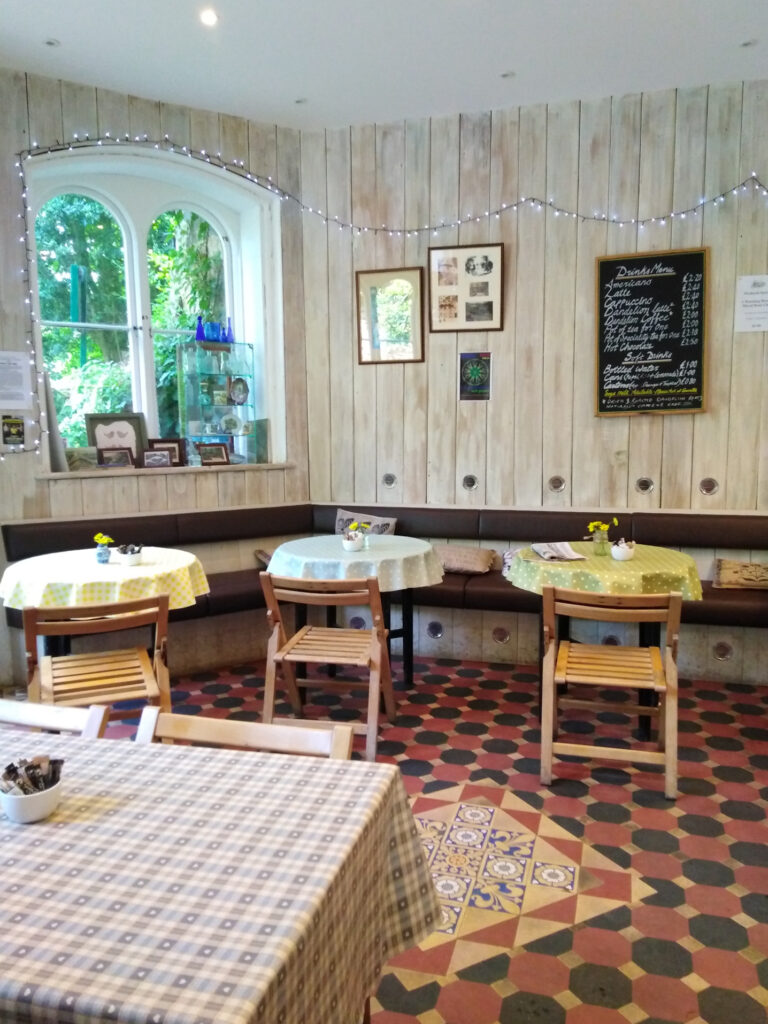 St Ann's Well Cafe interior with round tables, wooden chairs and a Victorian tiled floor. The walls are lined with white wooden panels and decorated with historical photos of the cafe and fairy lights.